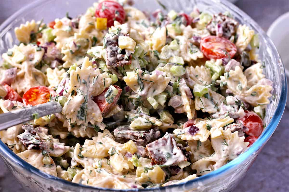 Pasta salad with bow tie pasta, vegan bacon, ranch dressing, chopped dill, and tomatoes is mixed in a glass bowl.