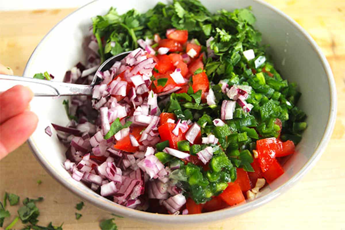 Diced red onions, diced tomatoes and jalapenos, and chopped cilantro are mixed with a spoon.