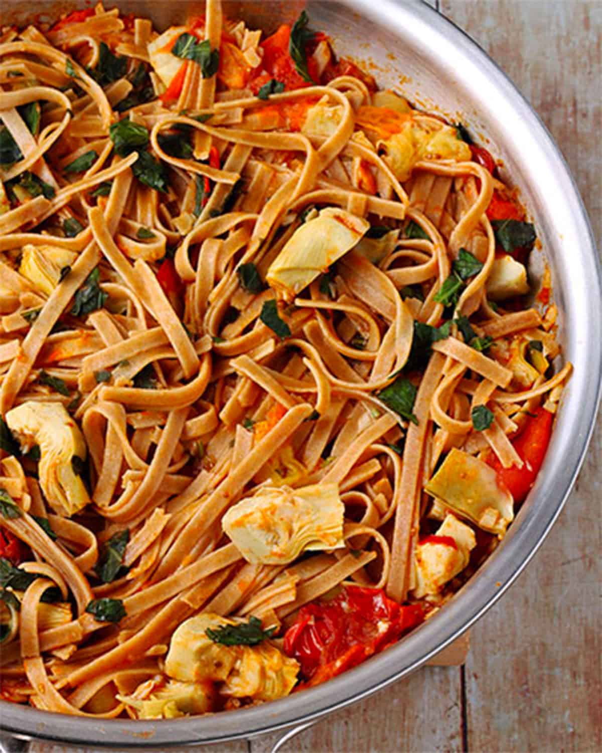 A pan with pasta, artichokes, cooked tomatoes, and fresh basil.