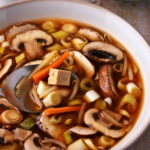 Hot and sour soup with mushrooms, scallions, carrots, bean sprouts, and tofu in a bowl with a spoon.