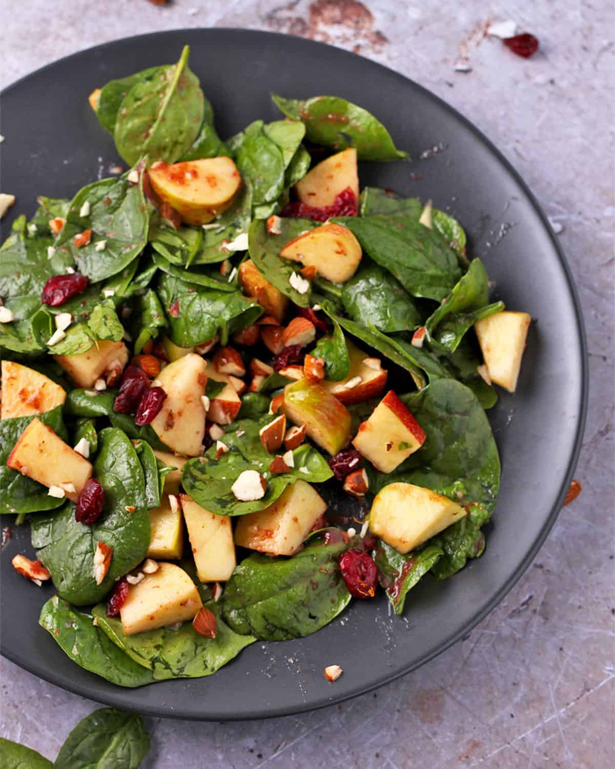 Spinach, apples, dried cranberries, and almonds with dressing on a black plate.