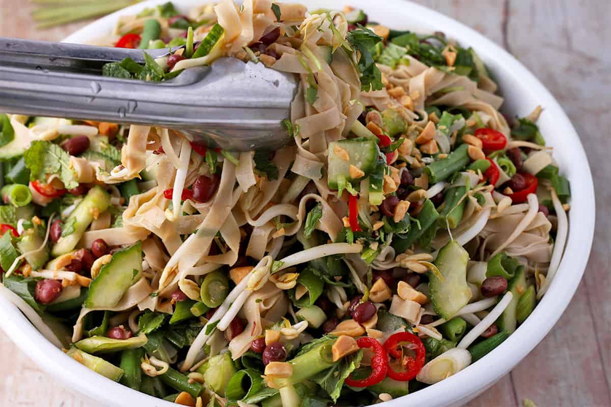 Rice noodles, a salad with herbs, green beans, bean sprouts, cucumber, green onions, sliced red chili, and peanuts is mixed with tongs.