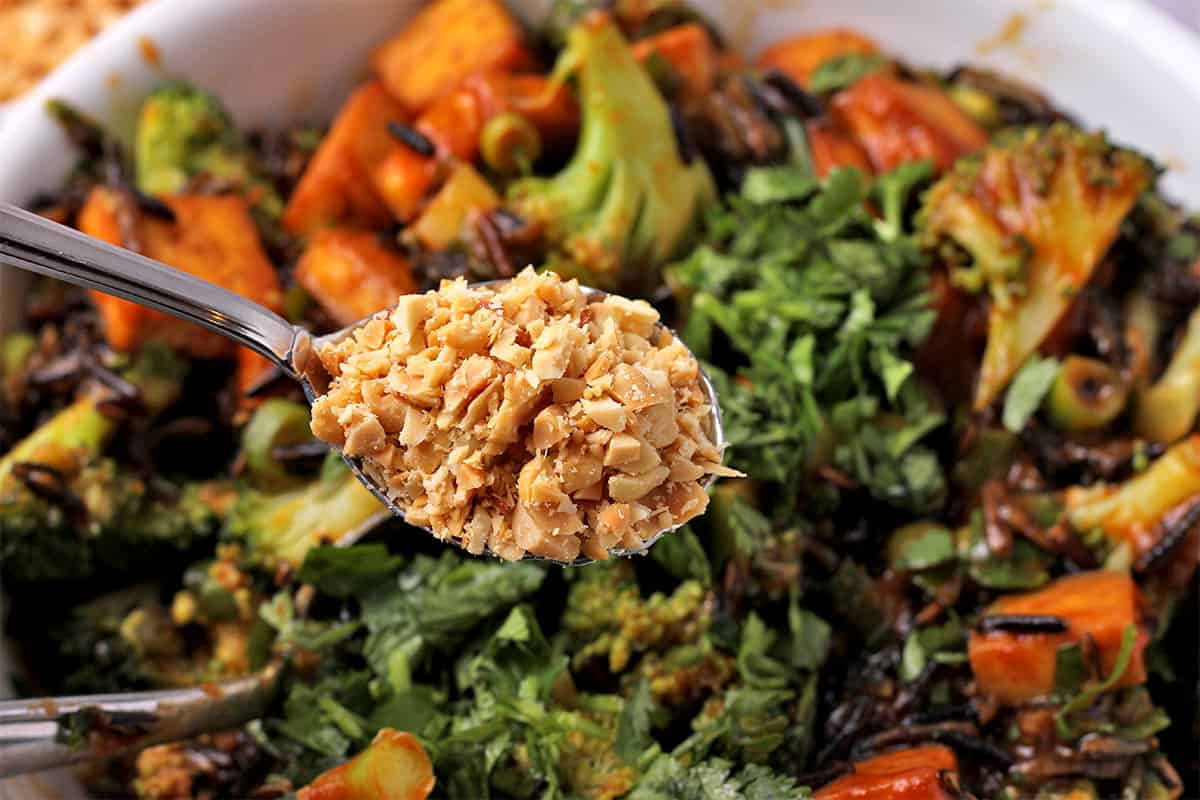 Chopped peanuts in a spoon held over a mixed salad with wild rice and broccoli.