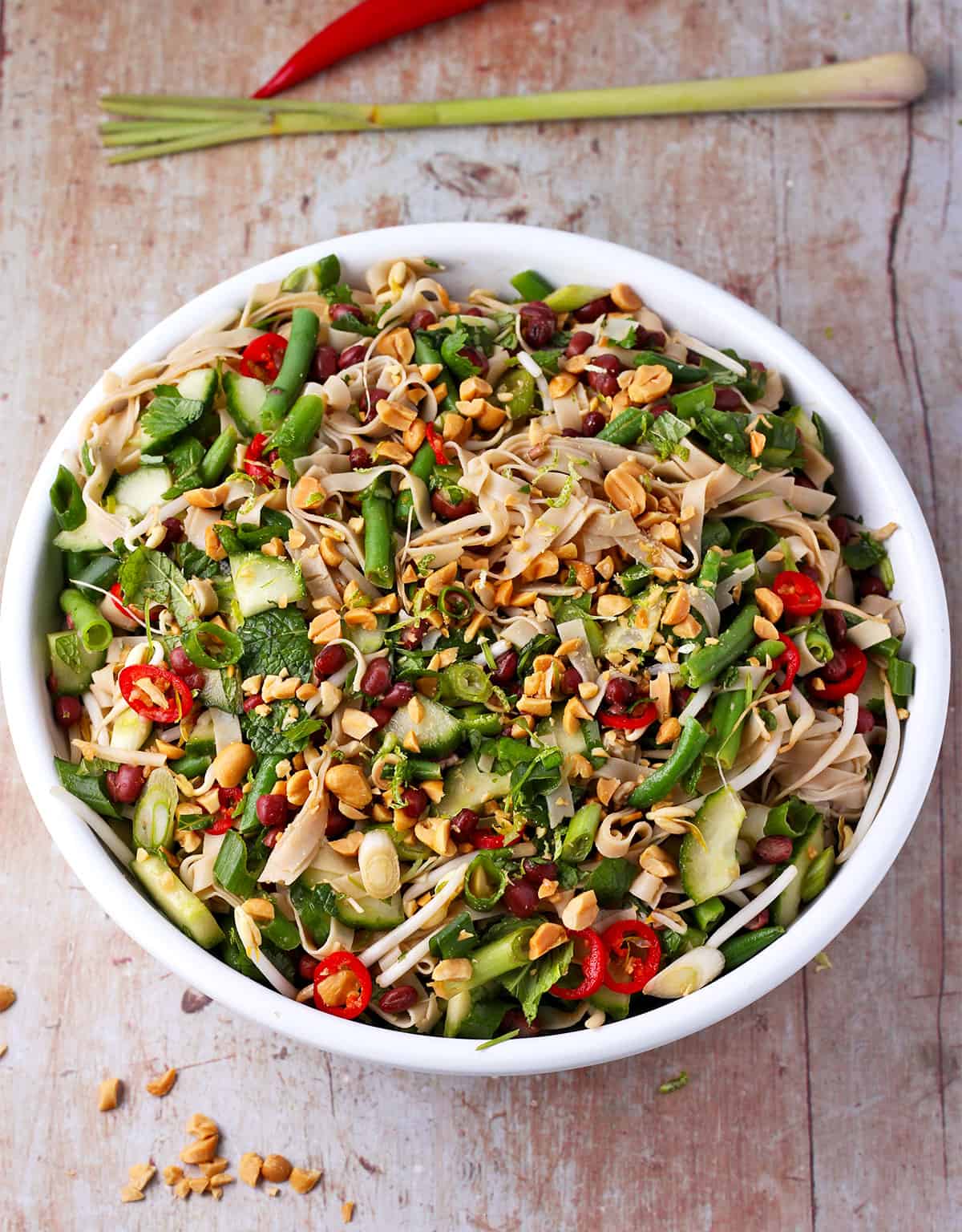 A white salad bowl is filled with green vegetables, adzuki beans, sliced red chilis, and chopped peanuts.