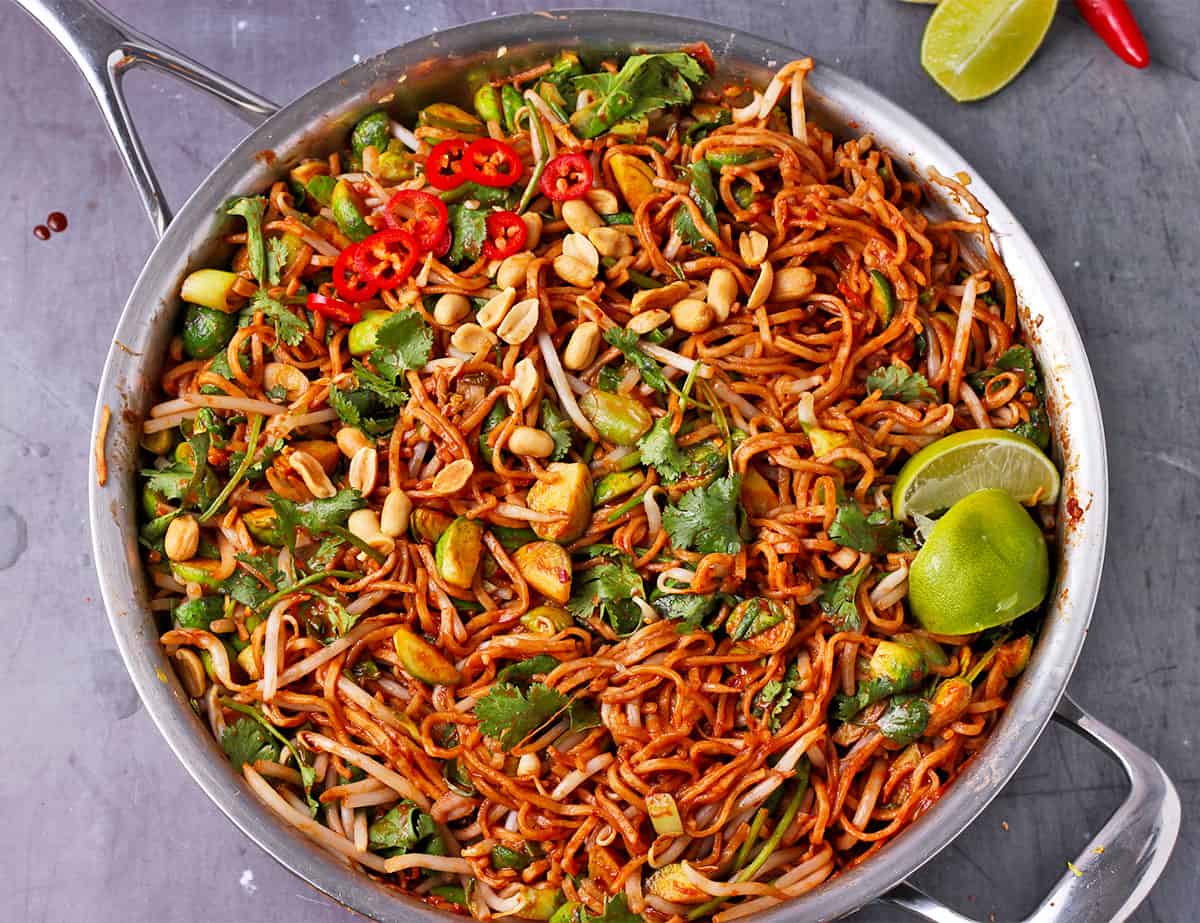 Noodles with bean sprouts, Brussels sprouts, cilantro, lime wedges, peanuts, and sliced red chili in a skillet.