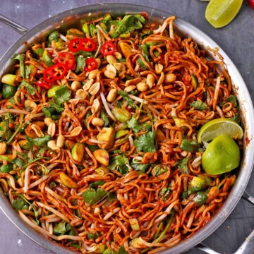Cooked noodles in sauce with Brussels sprouts, bean sprouts, cilantro, lime wedges, peanuts, and sliced red chili.