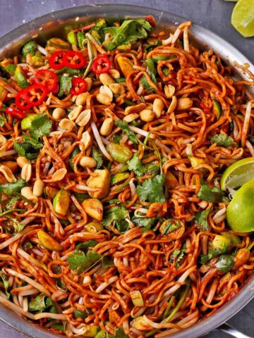 Cooked noodles in sauce with Brussels sprouts, bean sprouts, cilantro, lime wedges, peanuts, and sliced red chili.