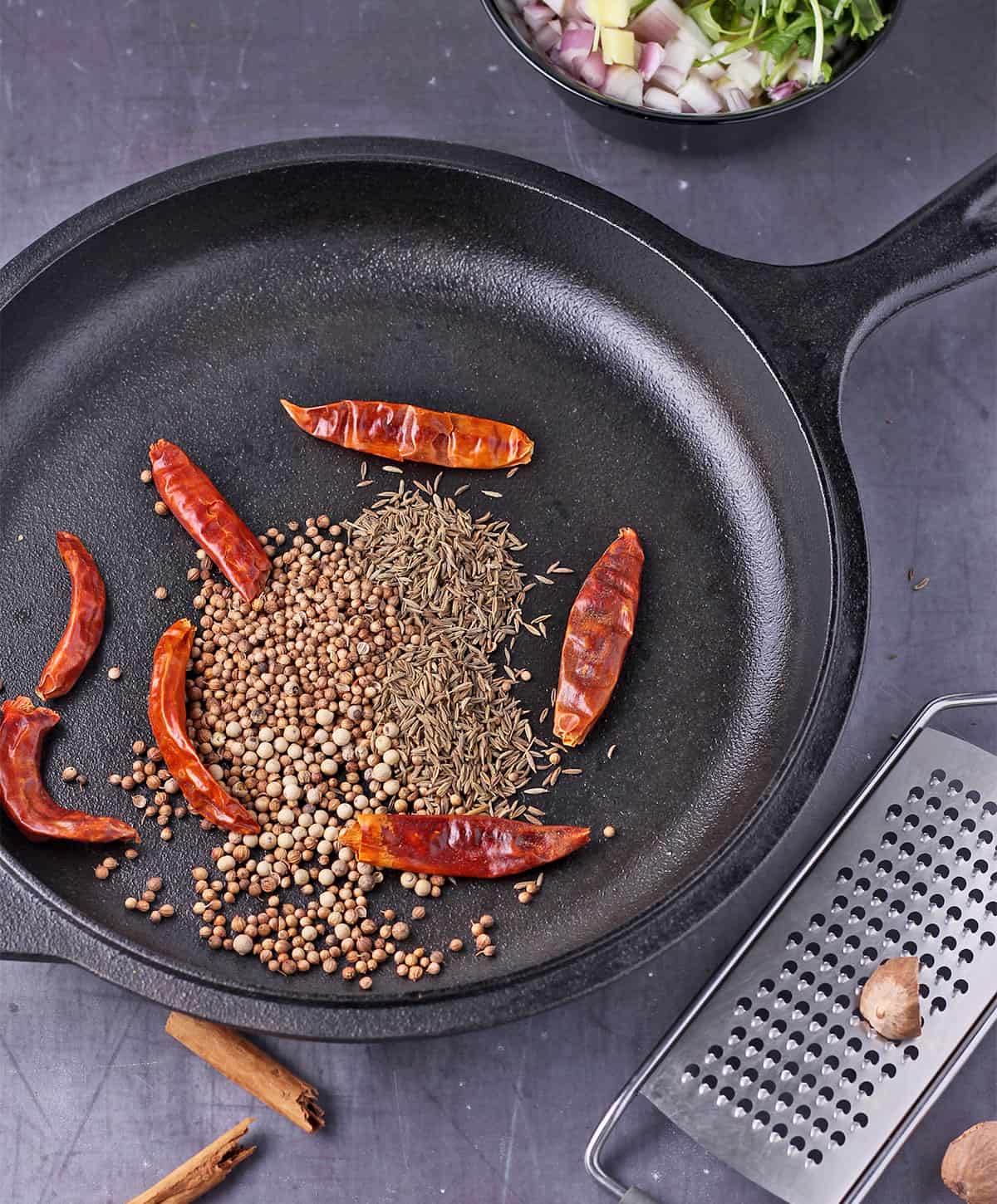 Dried red chilies, cumin seeds, coriander seeds, and cardamom seeds in a cast iron skillet with a nutmeg seed on a grater beside it.