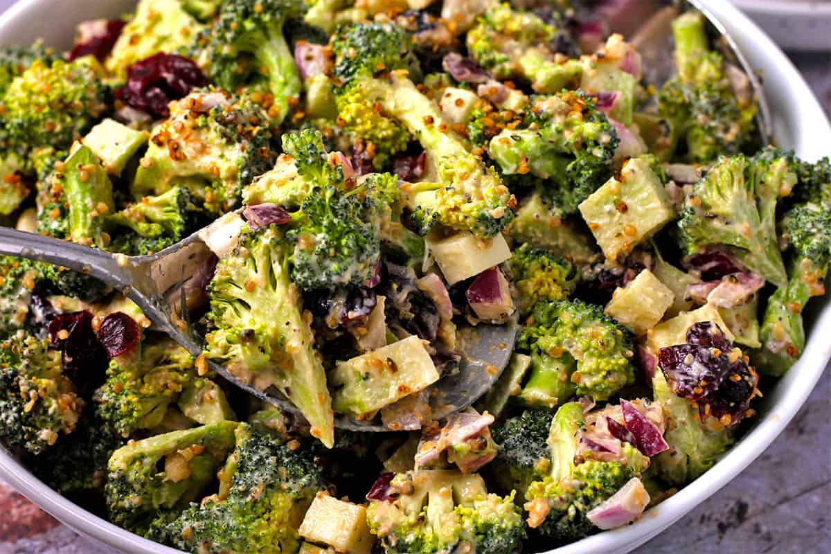 Broccoli salad is stirred with dressing, dried cranberries, red onions, and bacon bits.