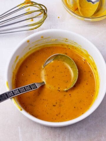 A ladle is in a white dish of vegan honey mustard dressing with black pepper.