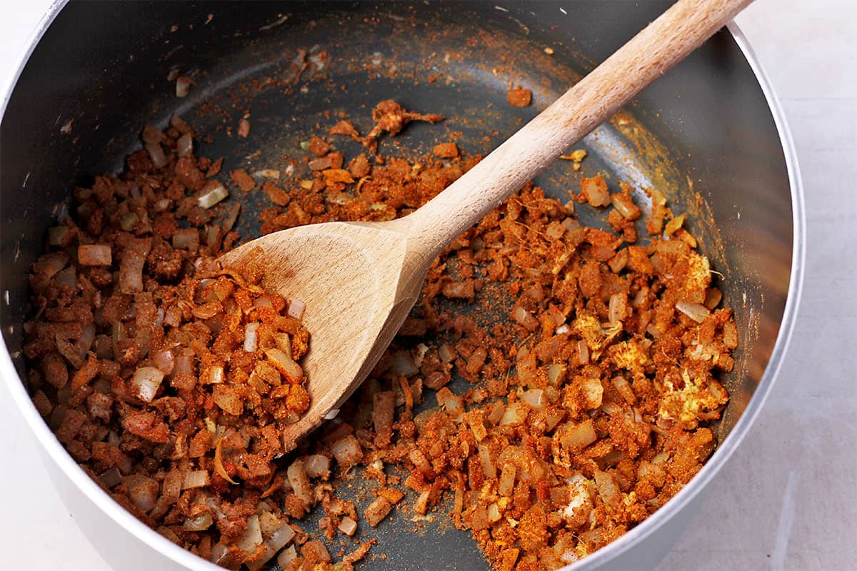 Spices and onions in a saucepan are stirred with a wooden spoon.