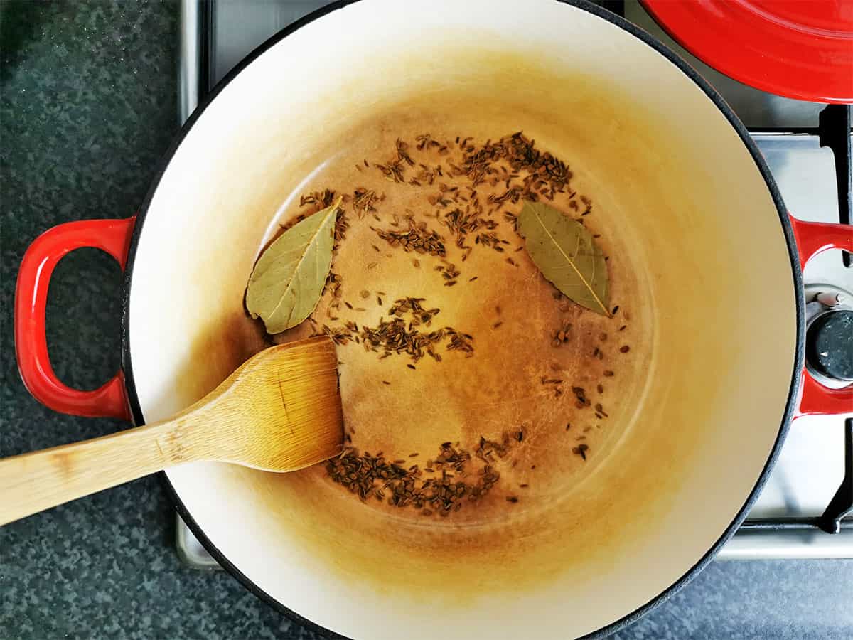 Whole spices and bay leaves in a pot are stirred with a flat wooden spoon.