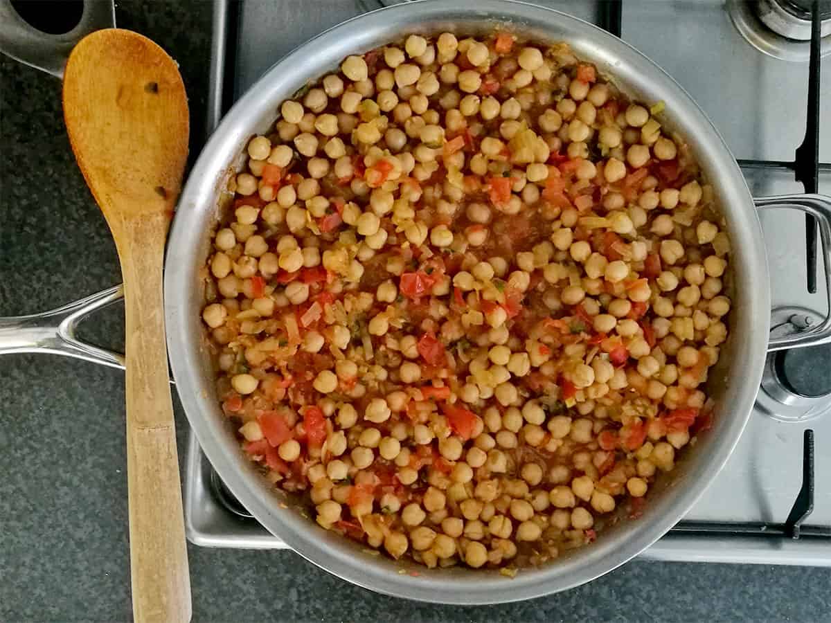 Chickpeas with chopped tomatoes in a frying pan with a wooden spoon on the side.