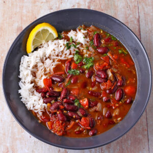 A bowl of kidney bean curry in a black bowl with white rice and a lemon slice.