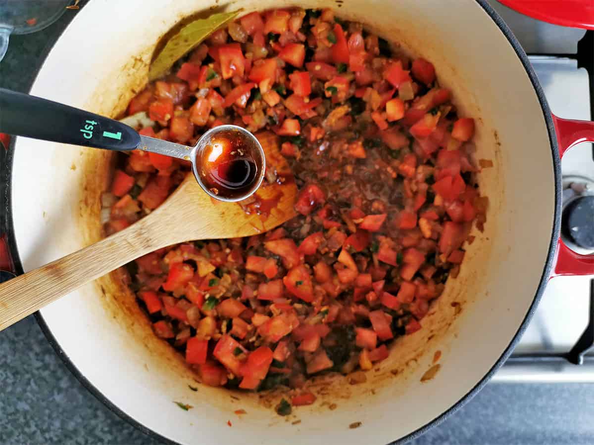 A teaspoon of date paste is held over a pot containing diced tomatoes, jalapenos, and onions.
