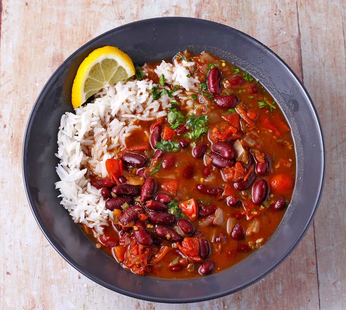 A black bowl of kidney bean curry with white rice and a lemon slice.