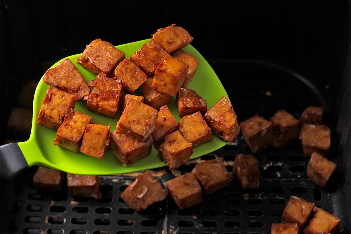 Crispy tofu cubes held over an air fryer basket in a green spatula.