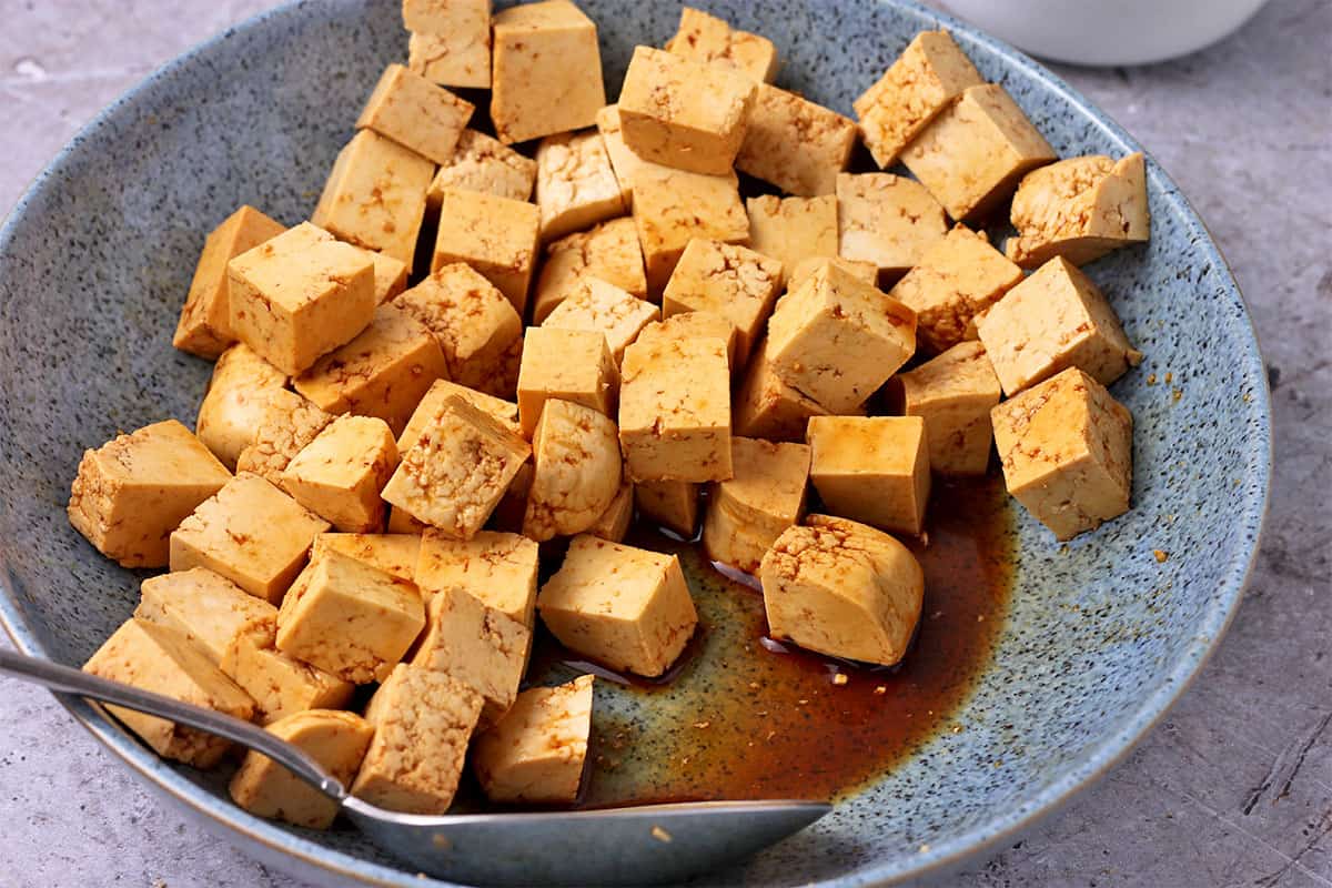 Tofu cubes are marinated in soy sauce in a blue bowl with a spoon.