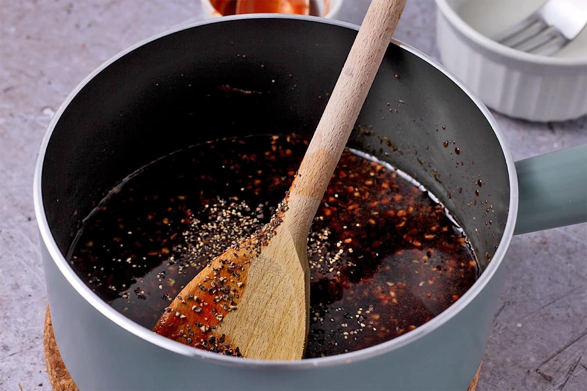 A saucepan with soy sauce, sriracha, and black pepper stirred with a wooden spoon.