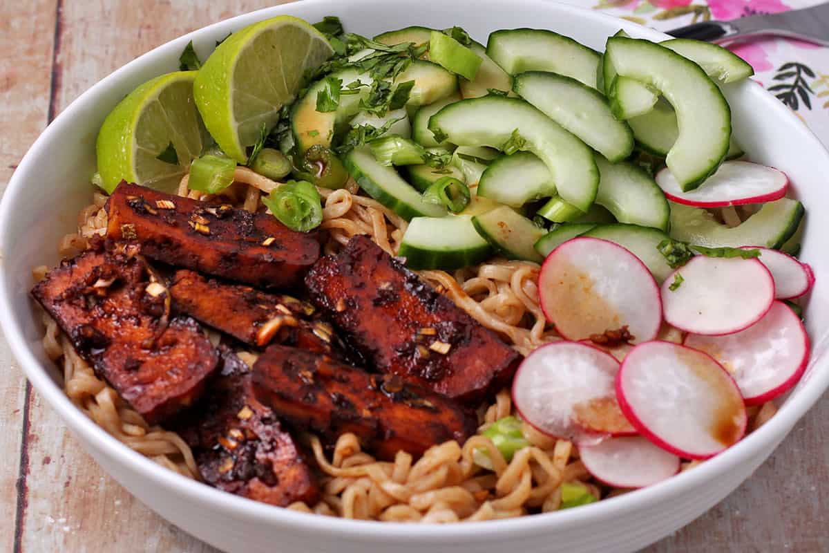 A bowl with baked tofu, noodles, sliced radishes and cucumbers, green onions, cilantro, and lime wedges.