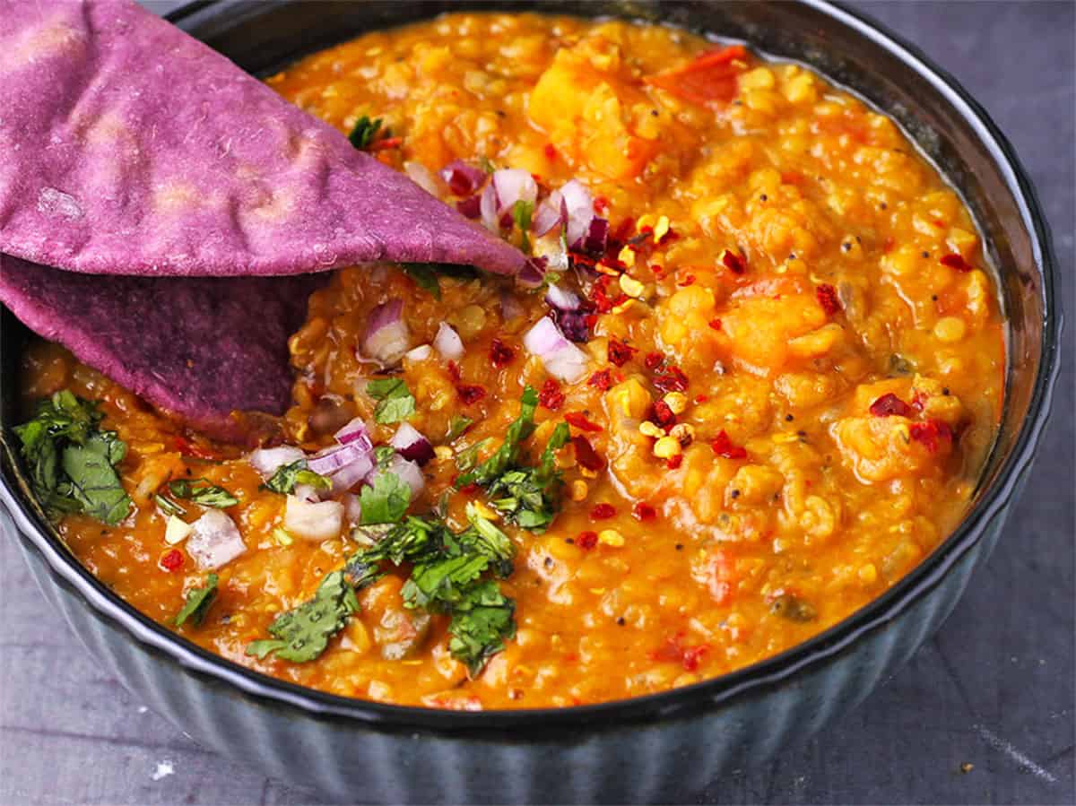 A bowl of red lentil dal with diced red onion, chopped cilantro, and a folded, purple flatbread.