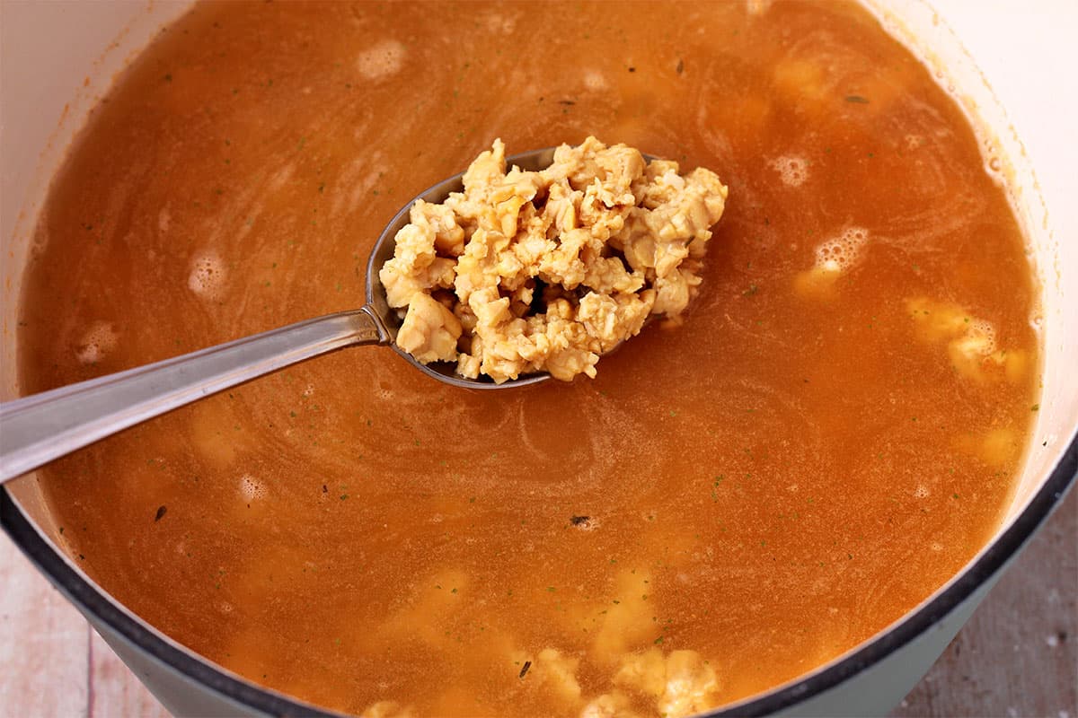 Simmered tempeh crumbles in a spoon over a pot of crumbles in vegetable broth.