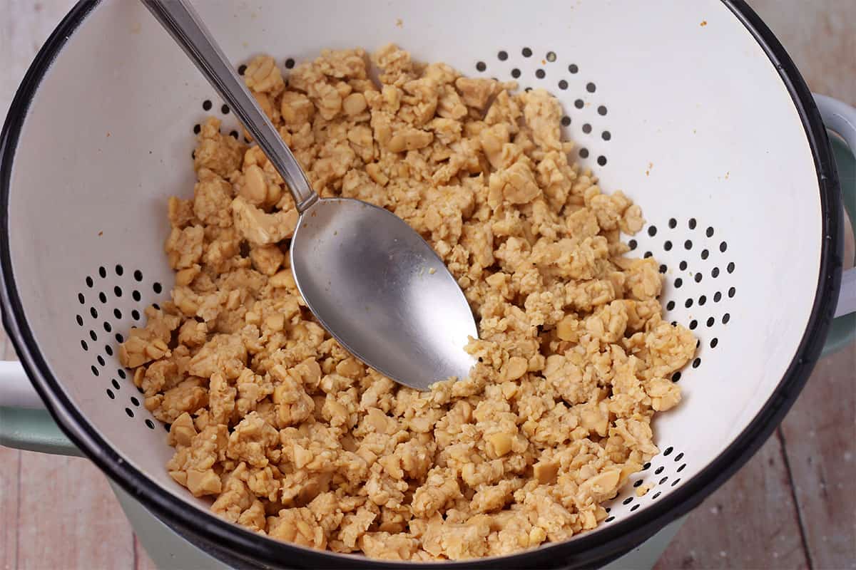 Tempeh crumbles in a colander are pressed with a spoon.
