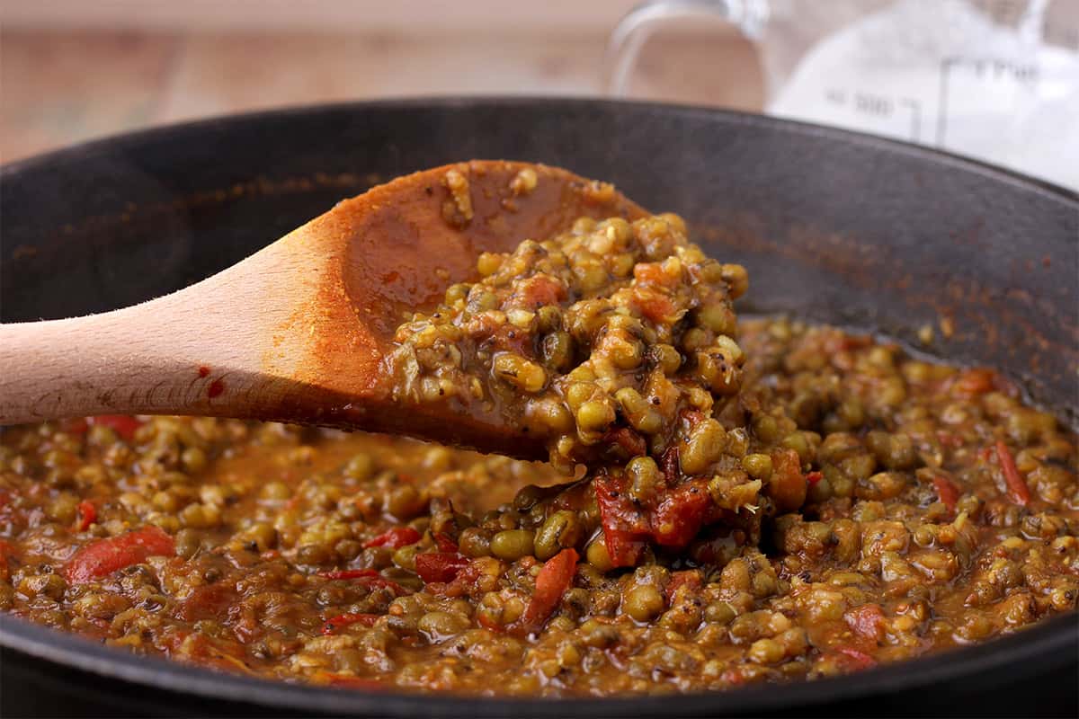 Cooked mung beans and tomatoes are stirred with a wooden spoon.