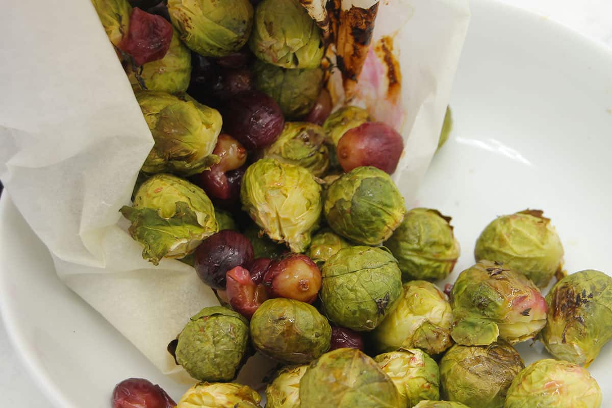 Roasted Brussels sprouts and grapes are transferred from parchment paper into a white bowl.