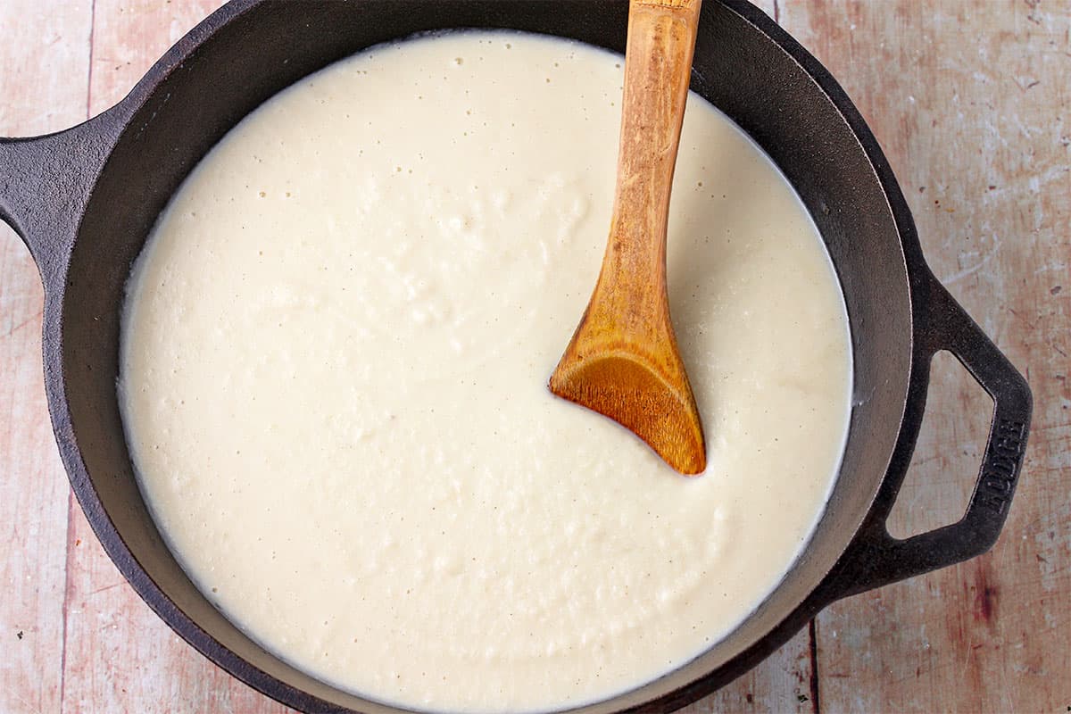 A black cast iron pan filled with vegan bechamel sauce with a wooden spoon in the sauce.