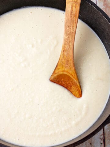 White bechamel sauce in a cast iron pan with a wooden spoon in the pan.