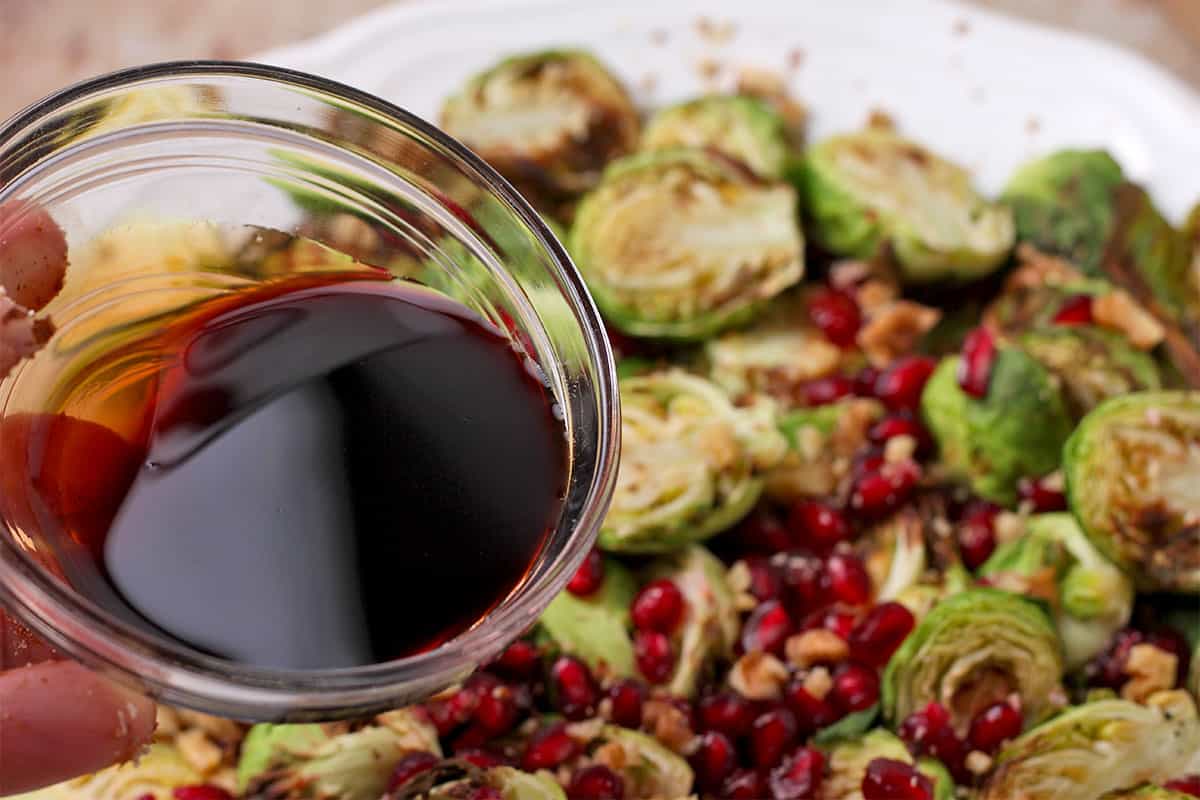 Pomegranate molasses in a small bowl is held over brussels sprouts with pomegranate.