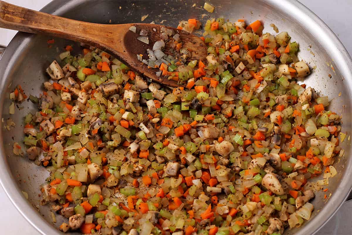Carrots, onions, celery, mushrooms, and spices are cooked in a pan and stirred with a wooden spoon.