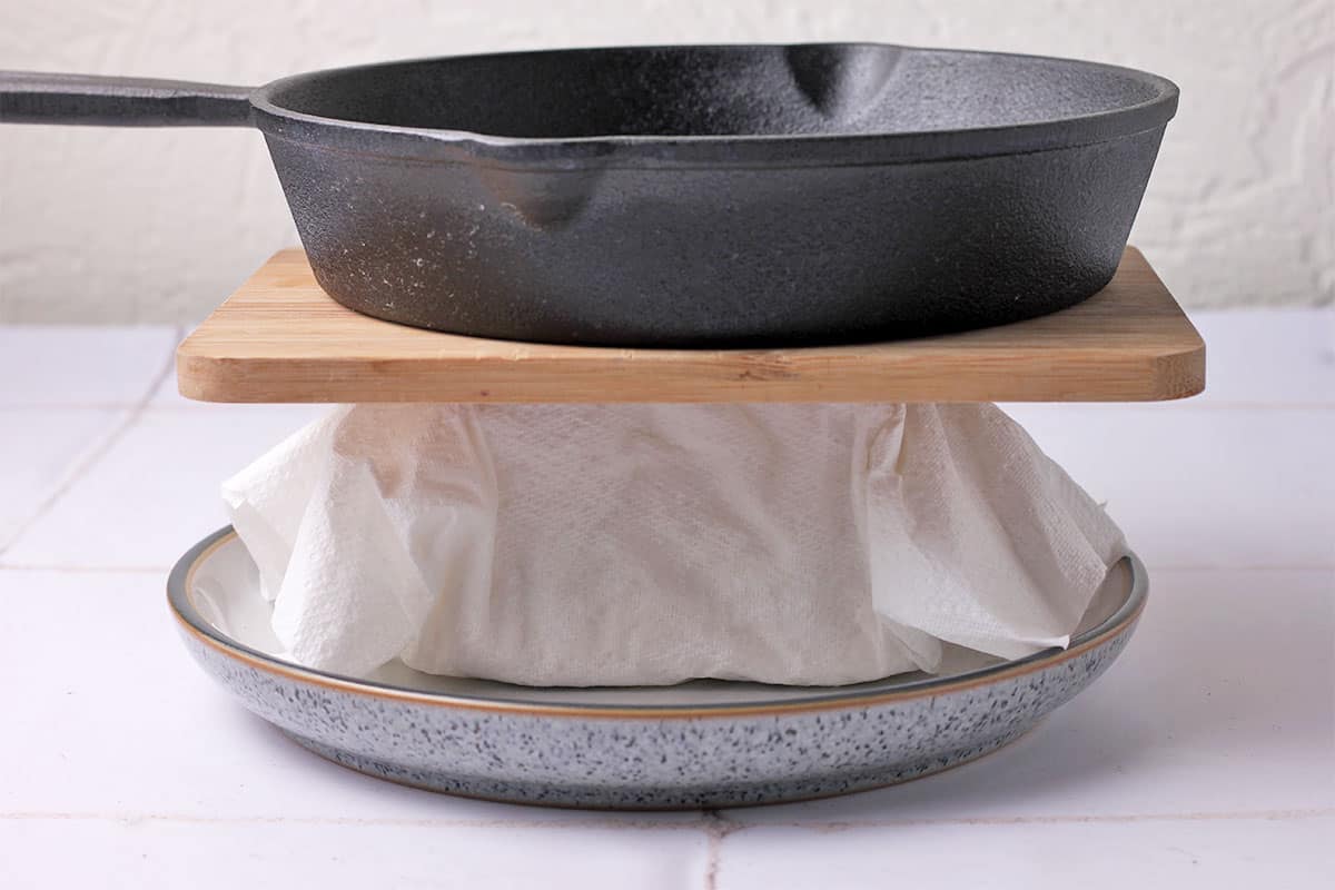 A block of tofu in paper towels is pressed between a plate and cutting board with cast iron skillet on top.