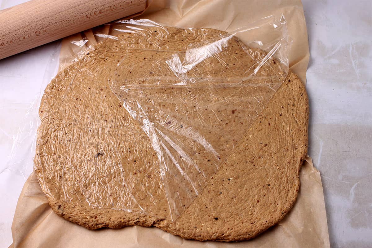 Seitan dough covered in cling film is rolled into a rectangle.