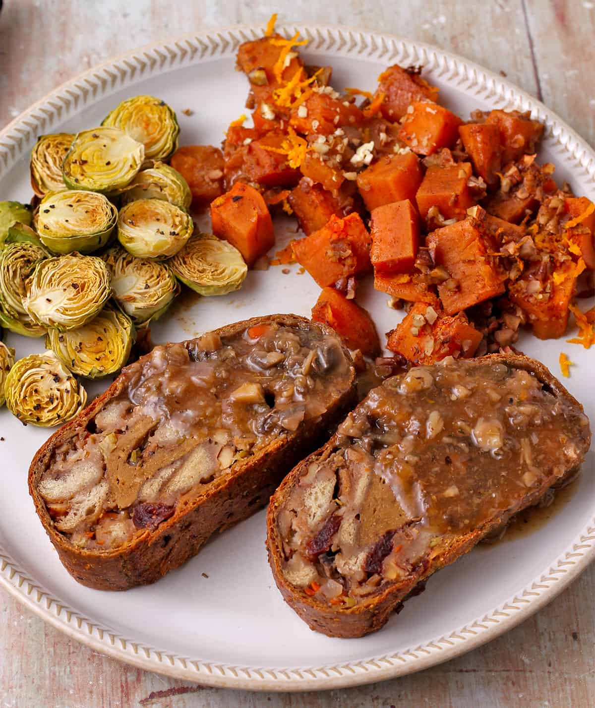 Stuffed seitan roast slices with gravy with diced sweet potatoes and roasted Brussels sprouts on a white plate.