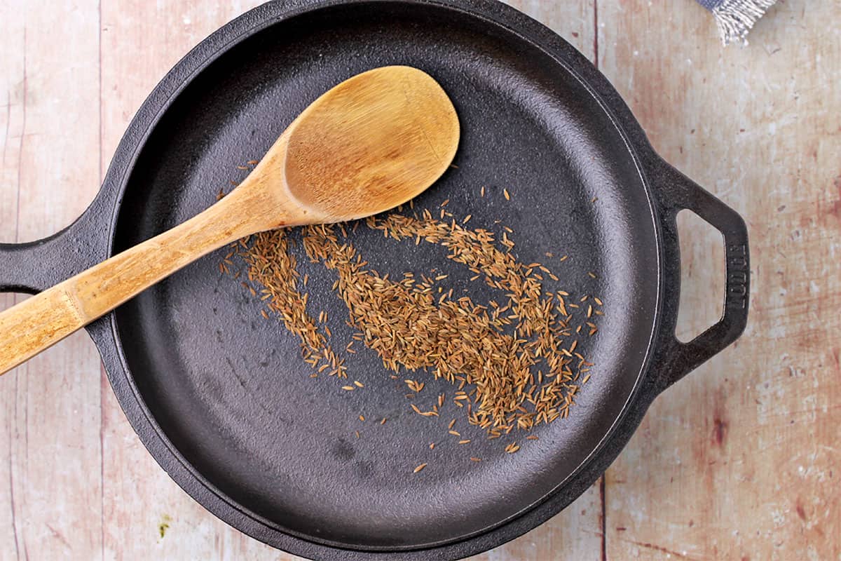 Cumin seeds and wooden spoon in a cast iron pan.