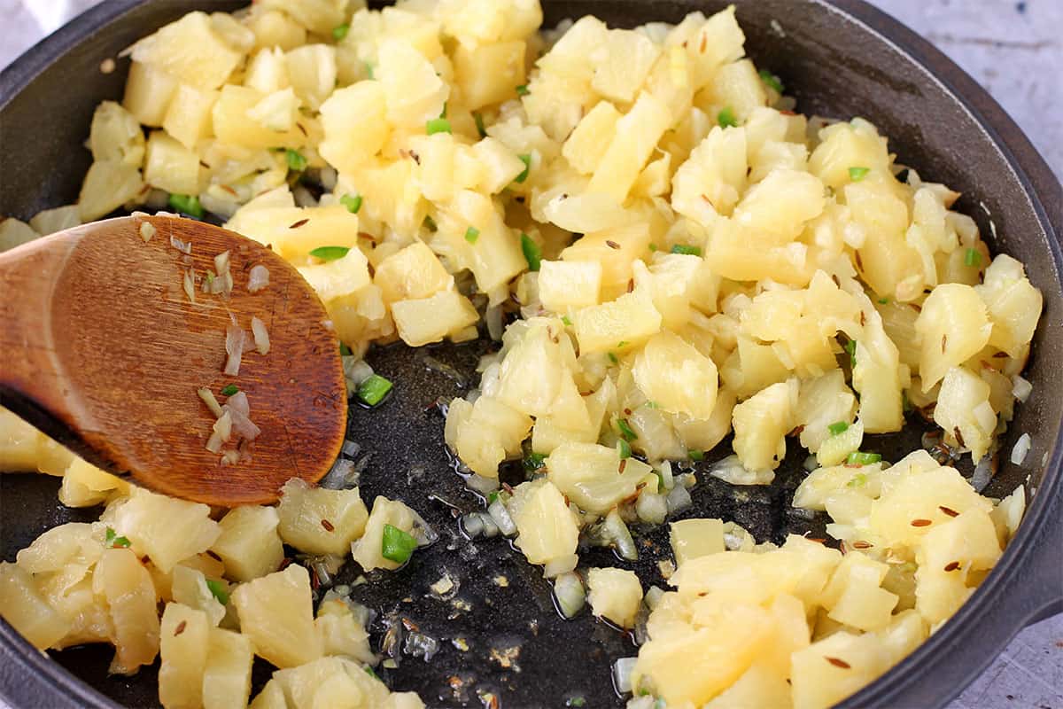 Pineapple pieces stirred with a wooden spoon in a cast iron pan.
