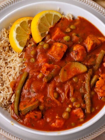 A white bowl with mixed vegetable and tomato curry with tofu, rice, and lemon wedges.