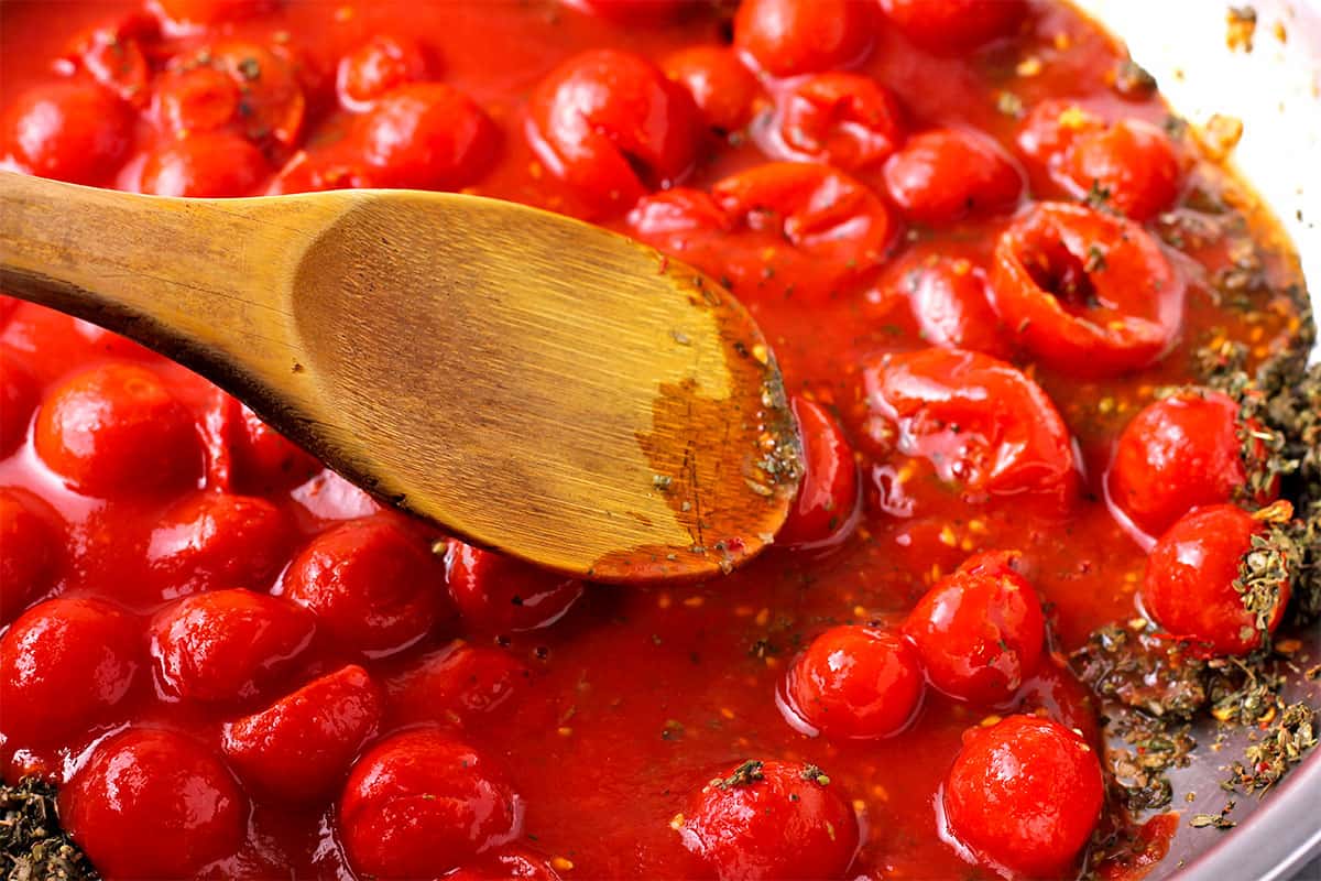 Canned tomatoes are stirred with a wooden spoon in a pan.