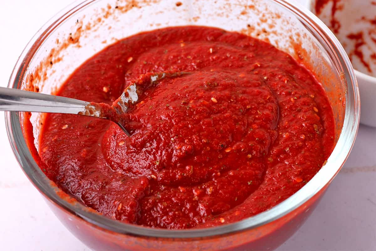 Spices are mixed into pizza sauce in a glass bowl.