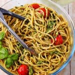 Cooked spaghetti with pesto, cherry tomatoes, and fresh basil in a glass bowl with tongs.