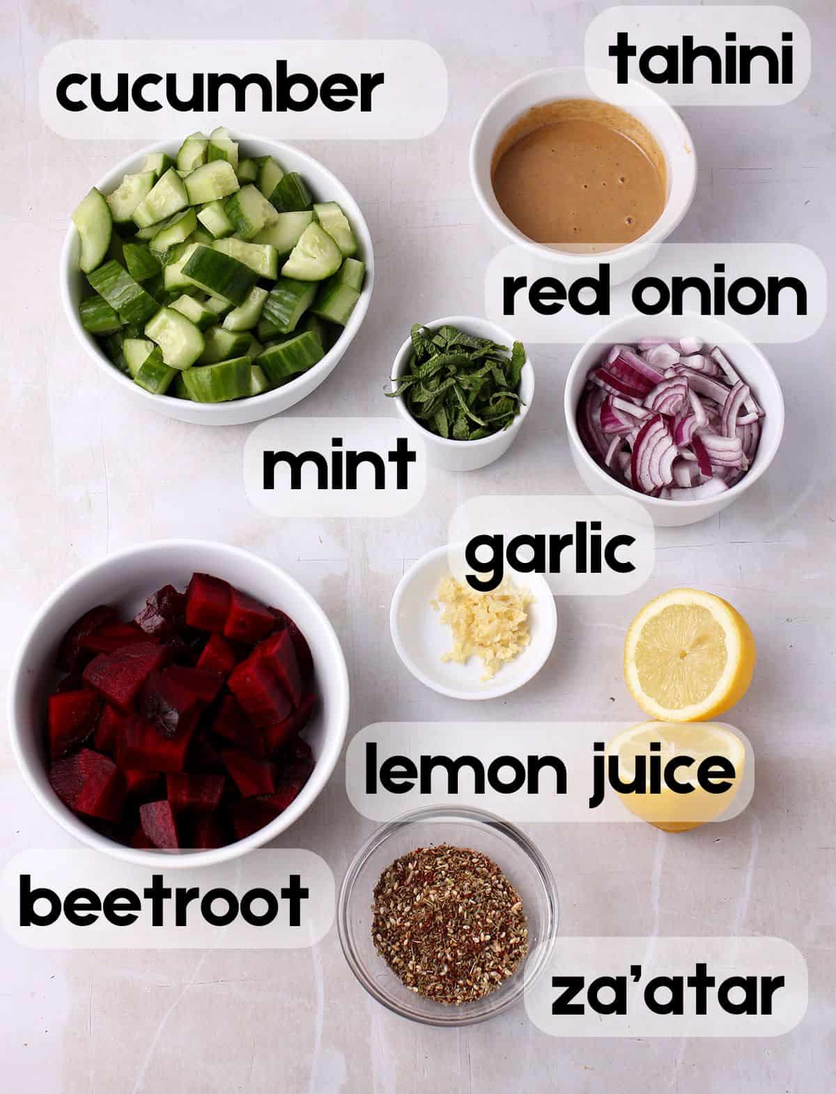 Cucumbers, tahini, beetroot, red onions, chopped mint, a cut lemon, garlic, and za'atar spice in small bowls with labels.