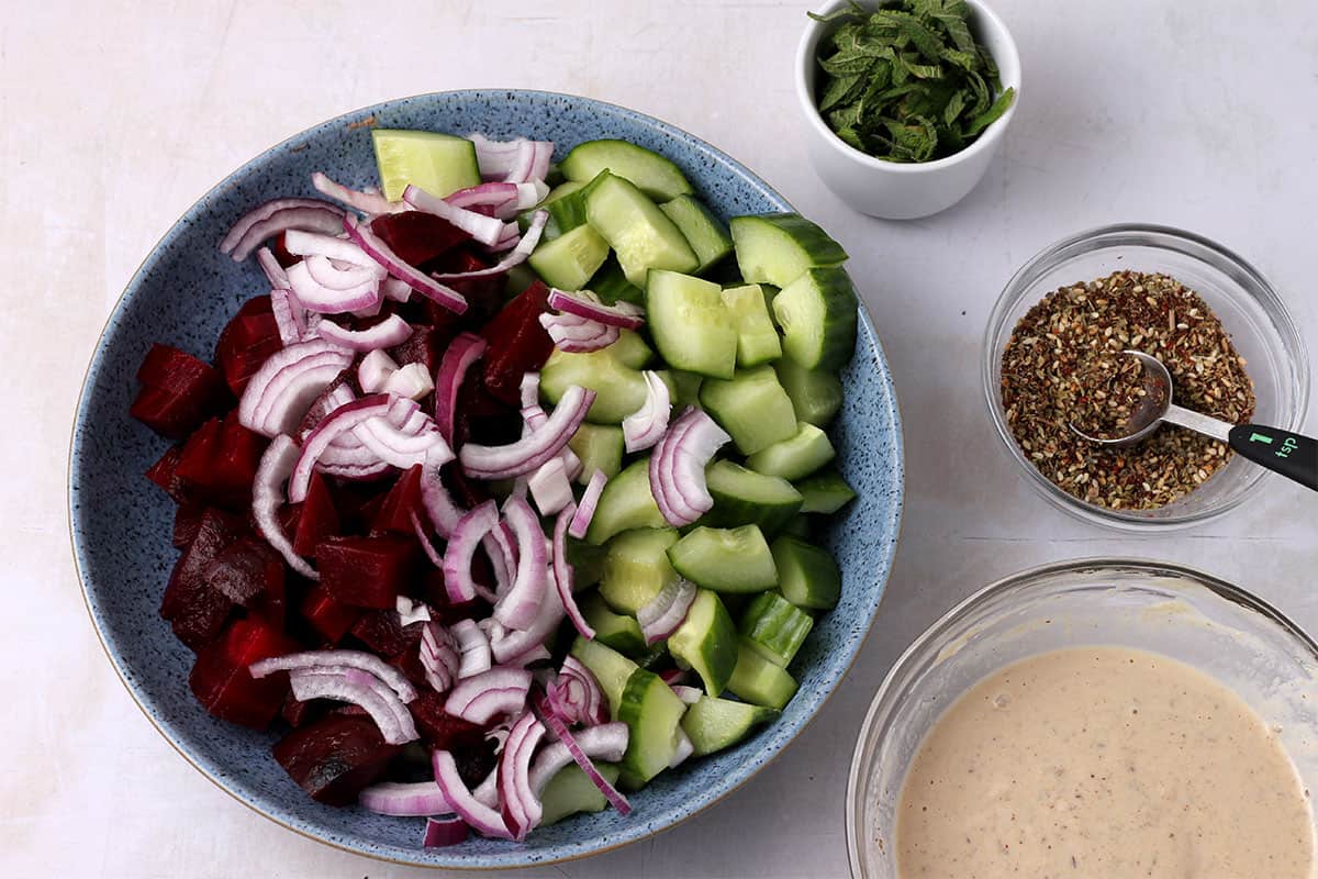Diced beets, cucumbers, and red onion in a bowl with another bowl of tahini dressing and a small bowl of za'atar seasoning.