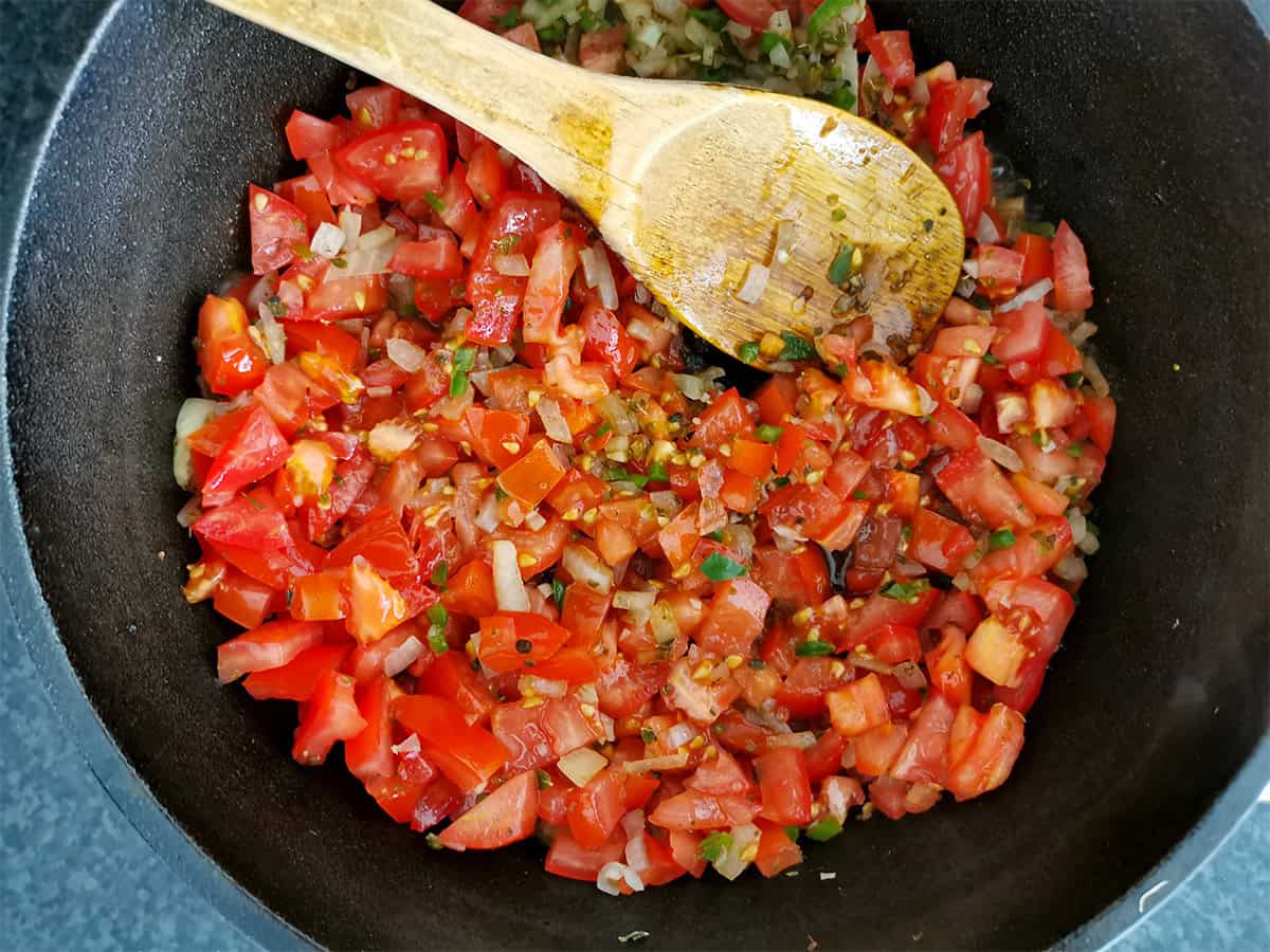 Chopped tomatoes, jalapenos, and onions are stirred in a cast iron pot.