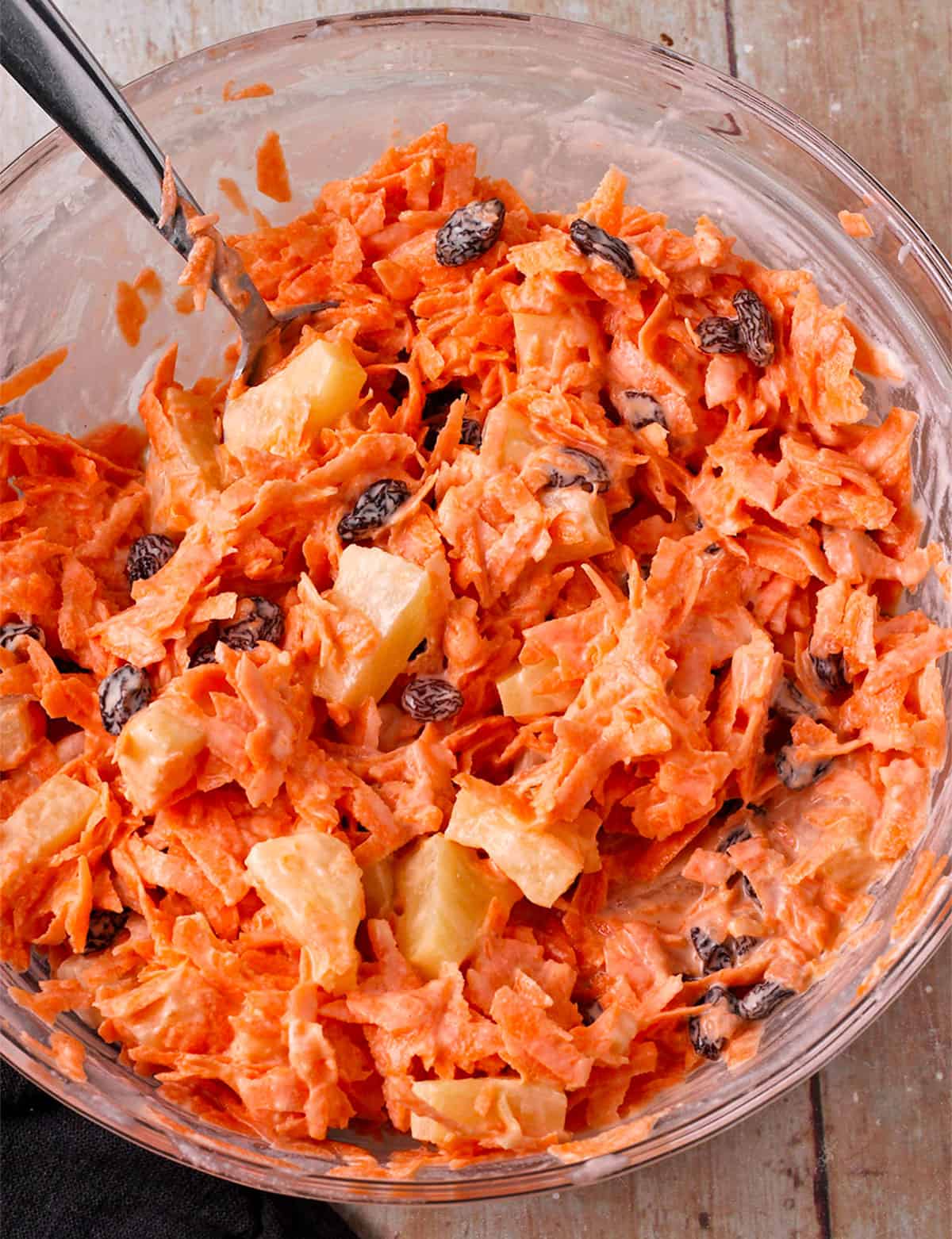 A glass bowl with shredded carrots, pineapple, raisins, and dressing.