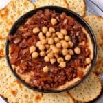 Hummus topped with balsamic caramelized onions and whole chickpeas in a bowl surrounded by crackers.