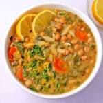 A bowl of lemon orzo chickpea soup with lemon wedges and chopped dill and parsley.