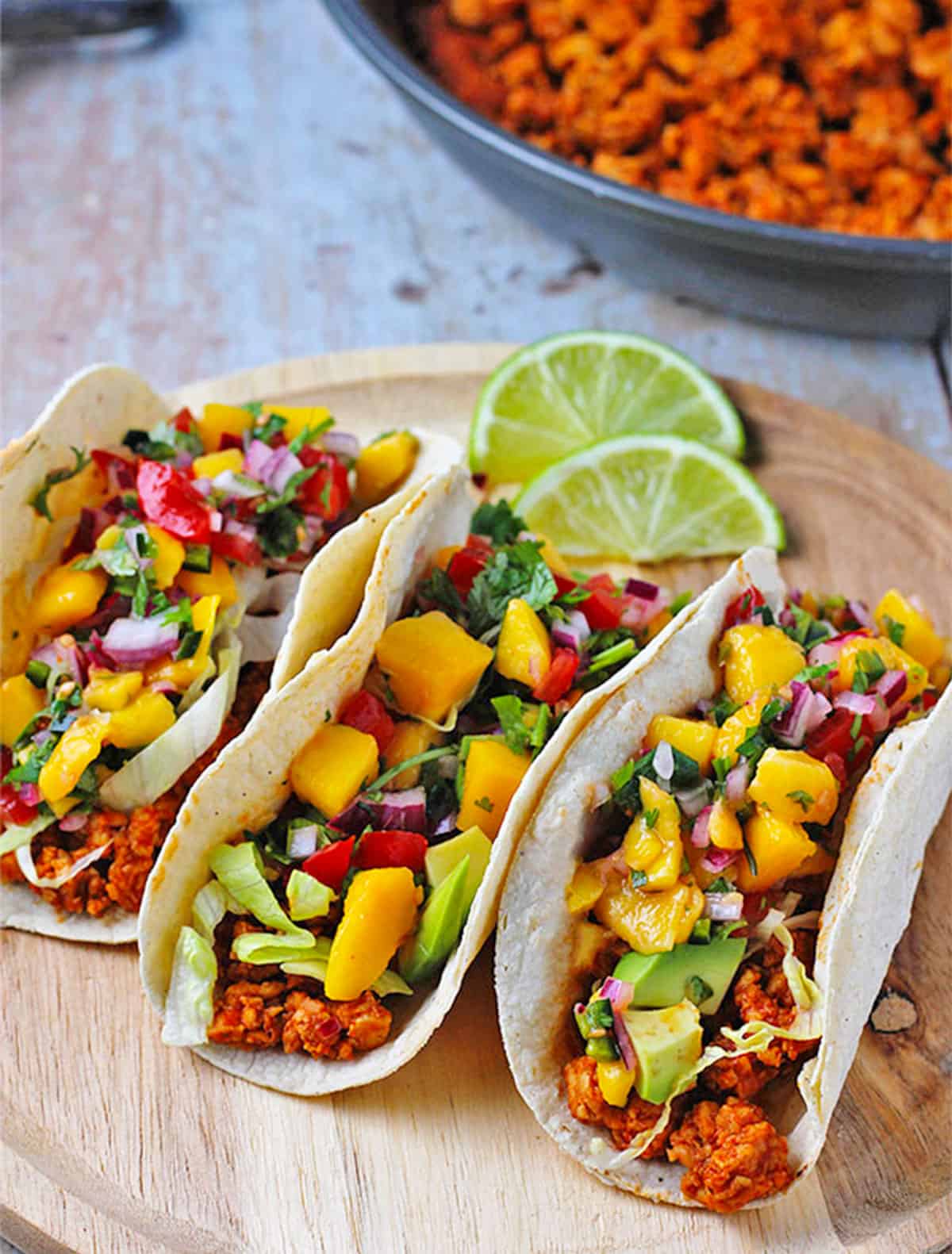 Tempeh asada tacos on a wooden plate with a pan of vegan carne asada in the background.