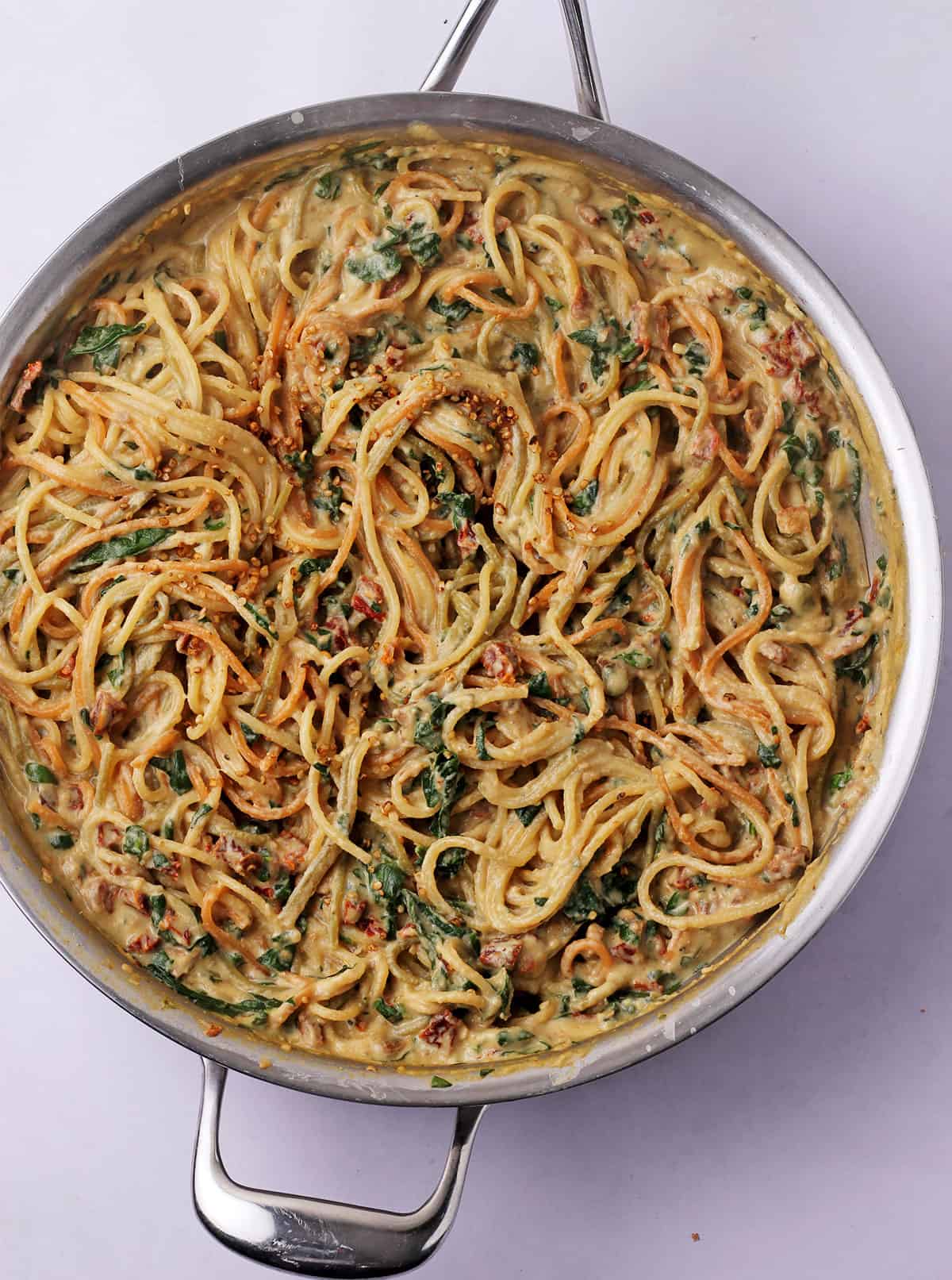 A frying pan filled with sundried tomato pasta with spinach and bacon bits.